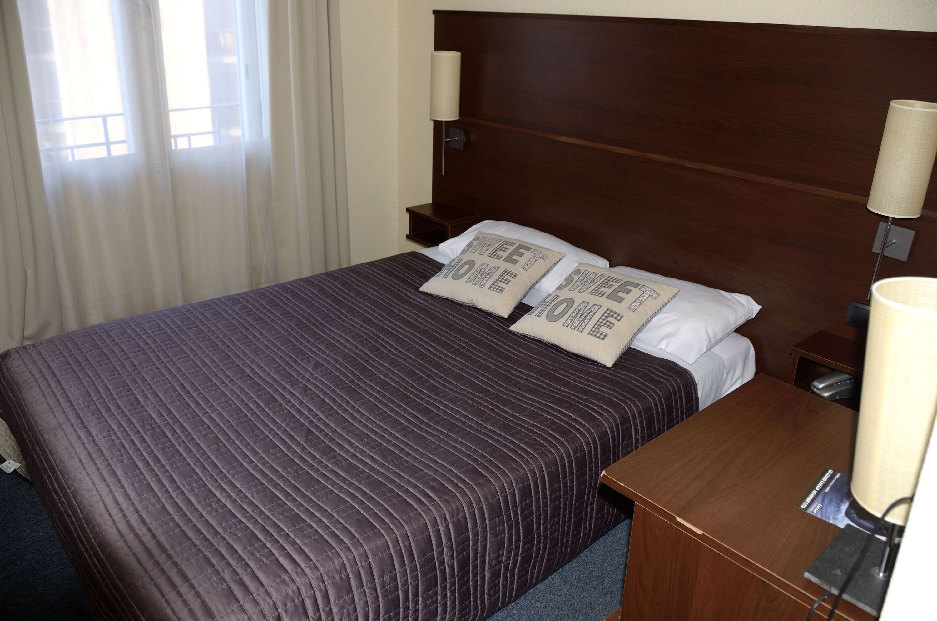 A neatly made bed with a dark quilt, two pillows with text, a wooden headboard, and a nightstand with a lamp.