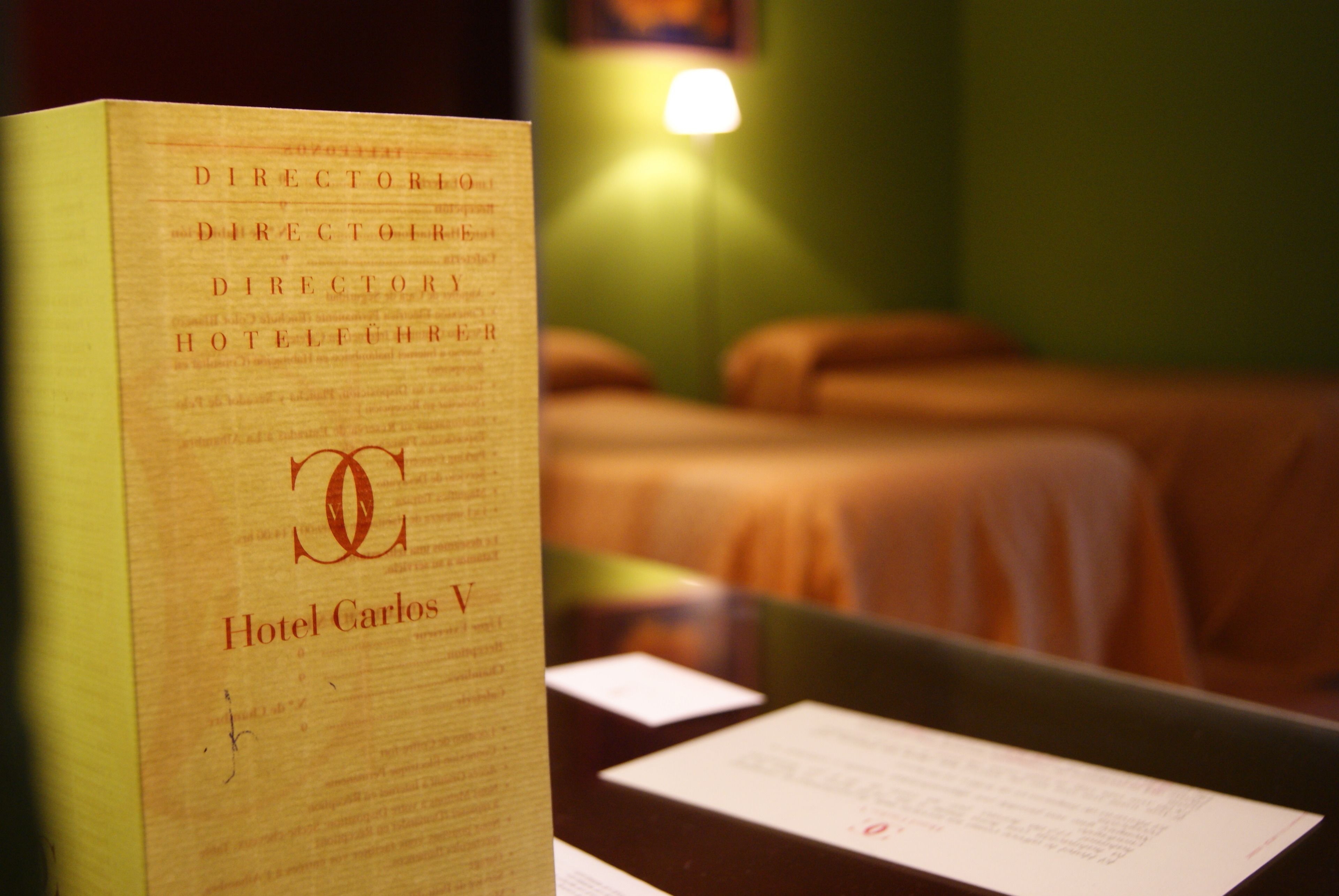 Un folleto de hotel titulado "Hotel Carlos V" que muestra una cama y una lámpara de noche al fondo.