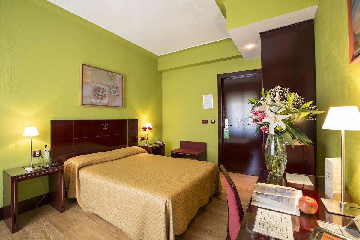 A hotel room with a bed, bedside tables, a desk with a lamp, a vase with flowers, and a door.