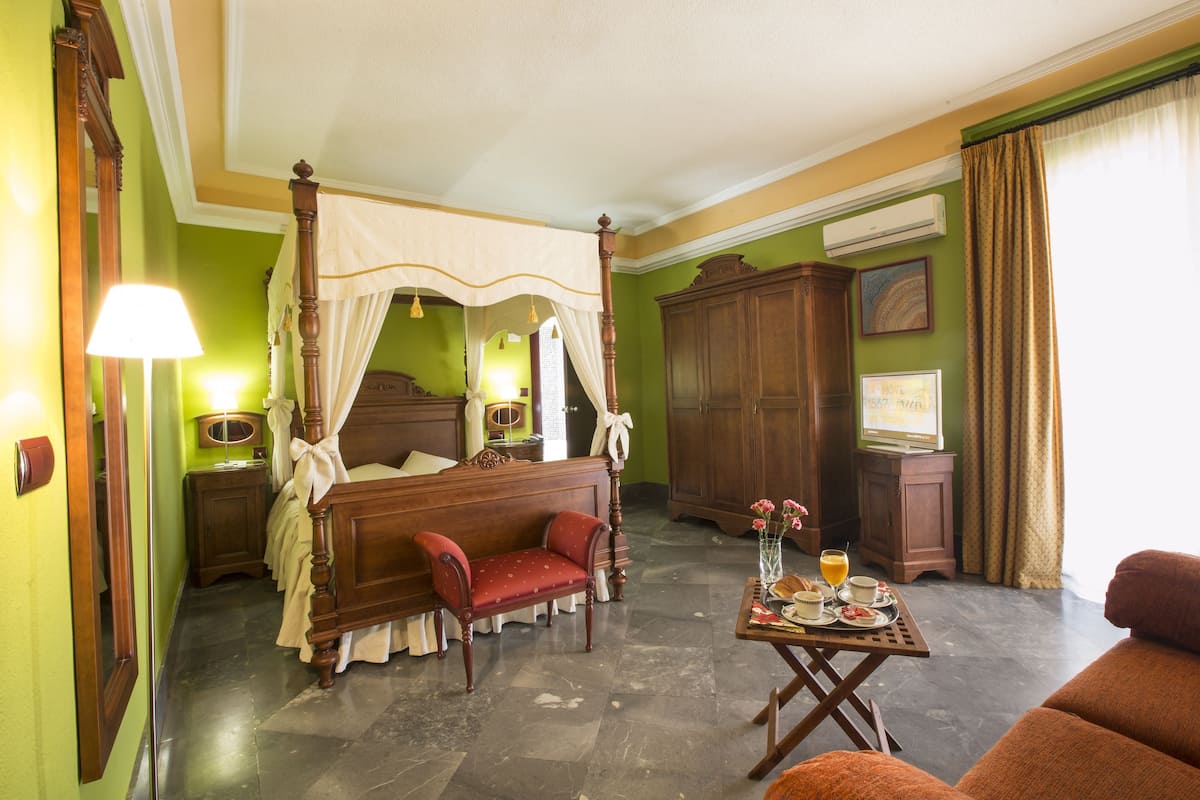 A room with a four-poster bed, a red armchair, a wooden cabinet, and a small table with a tray of breakfast items.