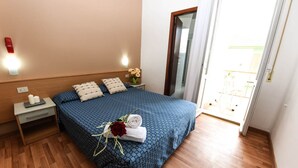 A neatly made bed with a blue patterned bedspread, a nightstand with a lamp, and a small table with a vase of flowers. - Hotel Miramare (Mondolfo)
