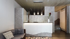 A modern reception area with a curved reception desk, a grey sofa with cushions, a patterned rug, and pendant lighting. - Hotel Miramare (Mondolfo)