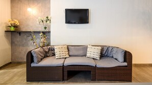 A wicker sofa with grey cushions, a flat-screen TV mounted on the wall, and a shelf with flowers. - Hotel Miramare (Mondolfo)
