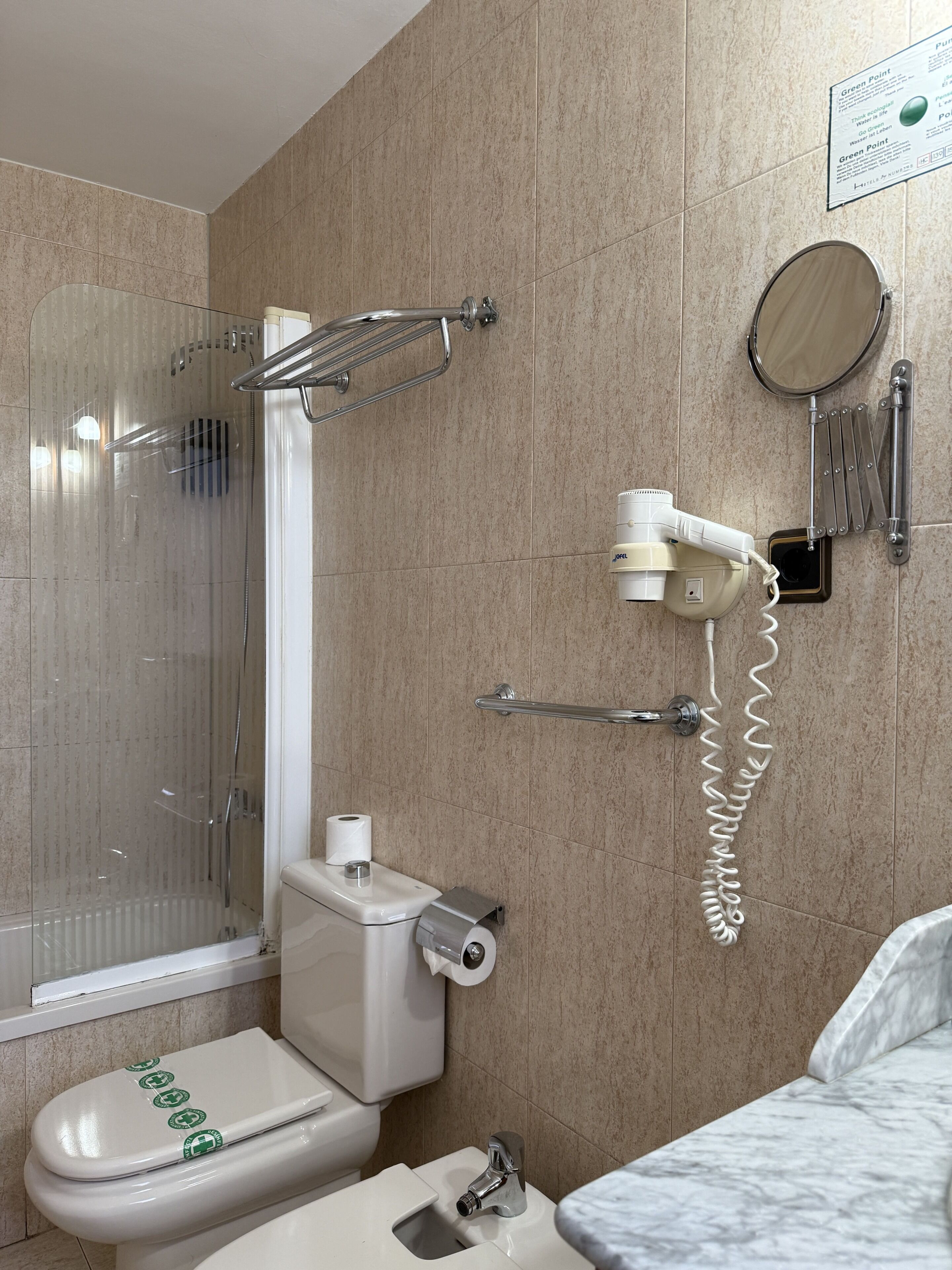 A bathroom with a glass-enclosed shower, a toilet with a paper towel dispenser, a hand dryer, and a towel rack.