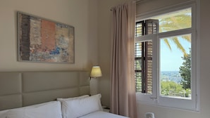 A hotel room with a bed, a painting on the wall, a lamp, a window with curtains, and a view of the city. - Hotel El Castell (Sant Boi de Llobregat)