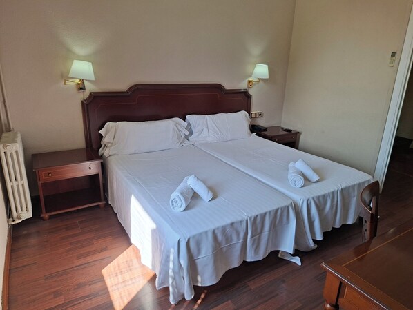 Un lit double avec du linge de lit blanc, deux tables de chevet avec des lampes, une tête de lit en bois et un radiateur.