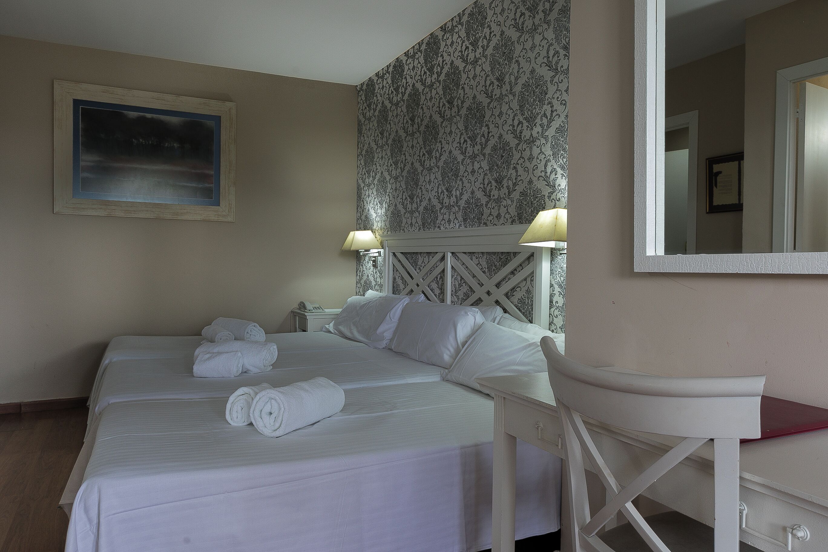 A hotel room with two beds, a desk, a chair, a mirror, and a patterned wallpaper.