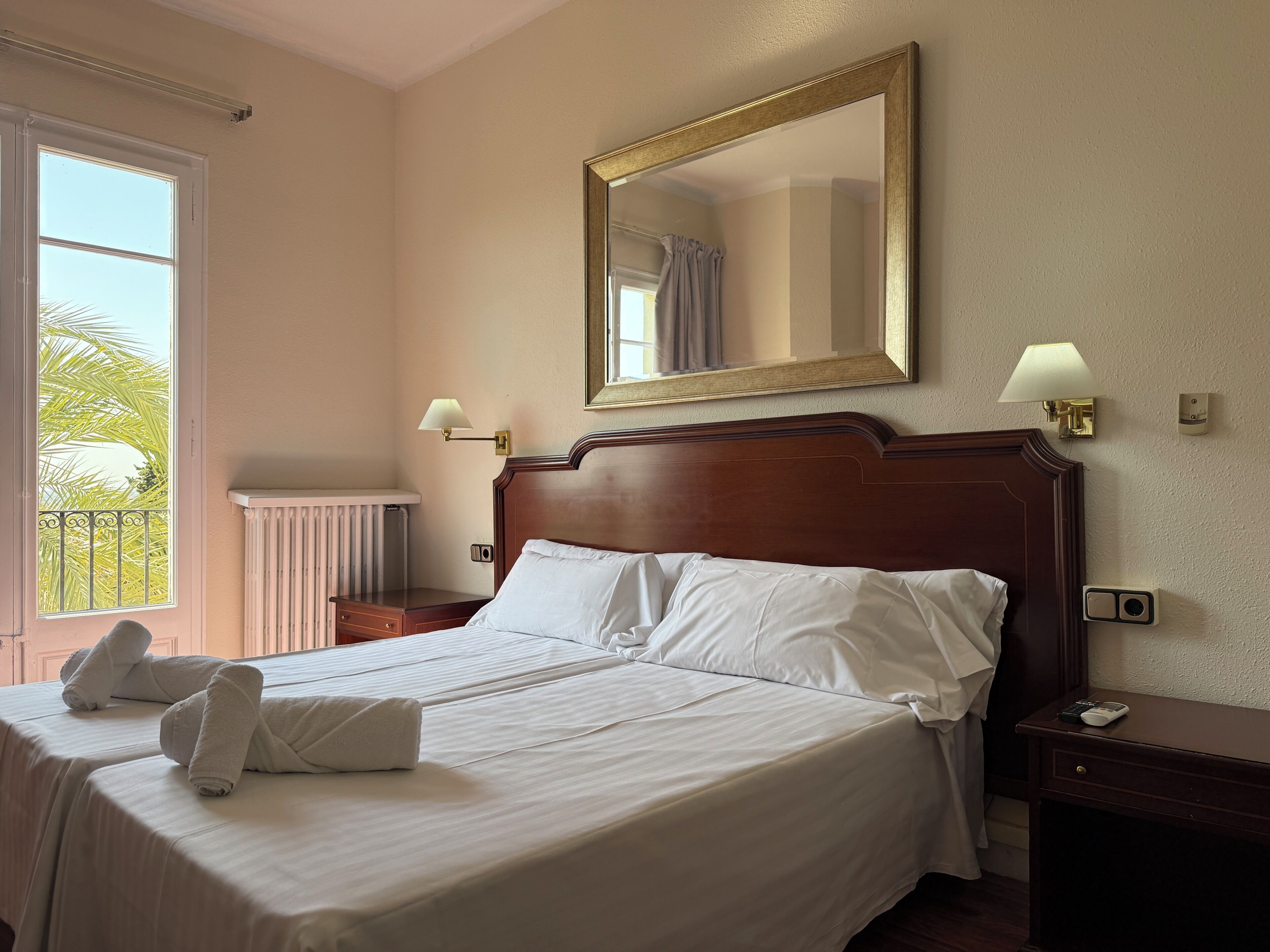 Una cama bien hecha con sábanas y toallas blancas, un espejo grande encima y una ventana con vistas a palmeras.