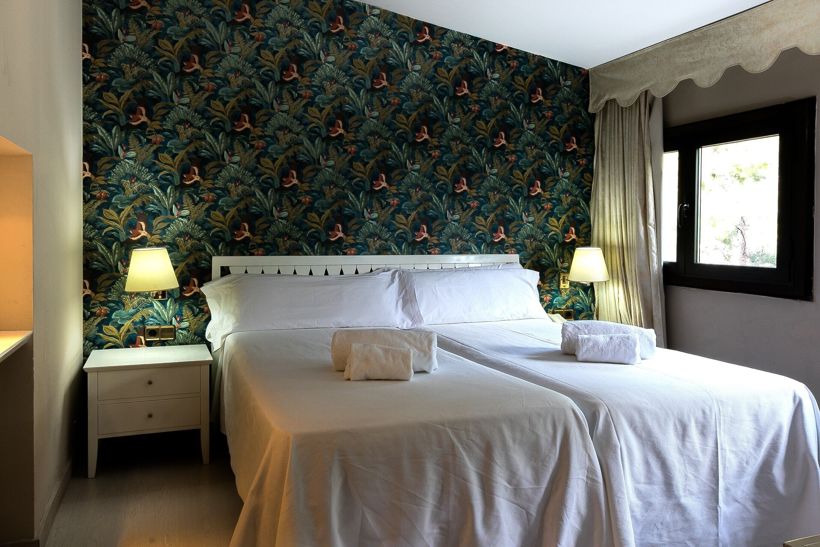 Una habitación de hotel con una cama, dos mesitas de noche con lámparas, una ventana con cortinas y un papel tapiz estampado.