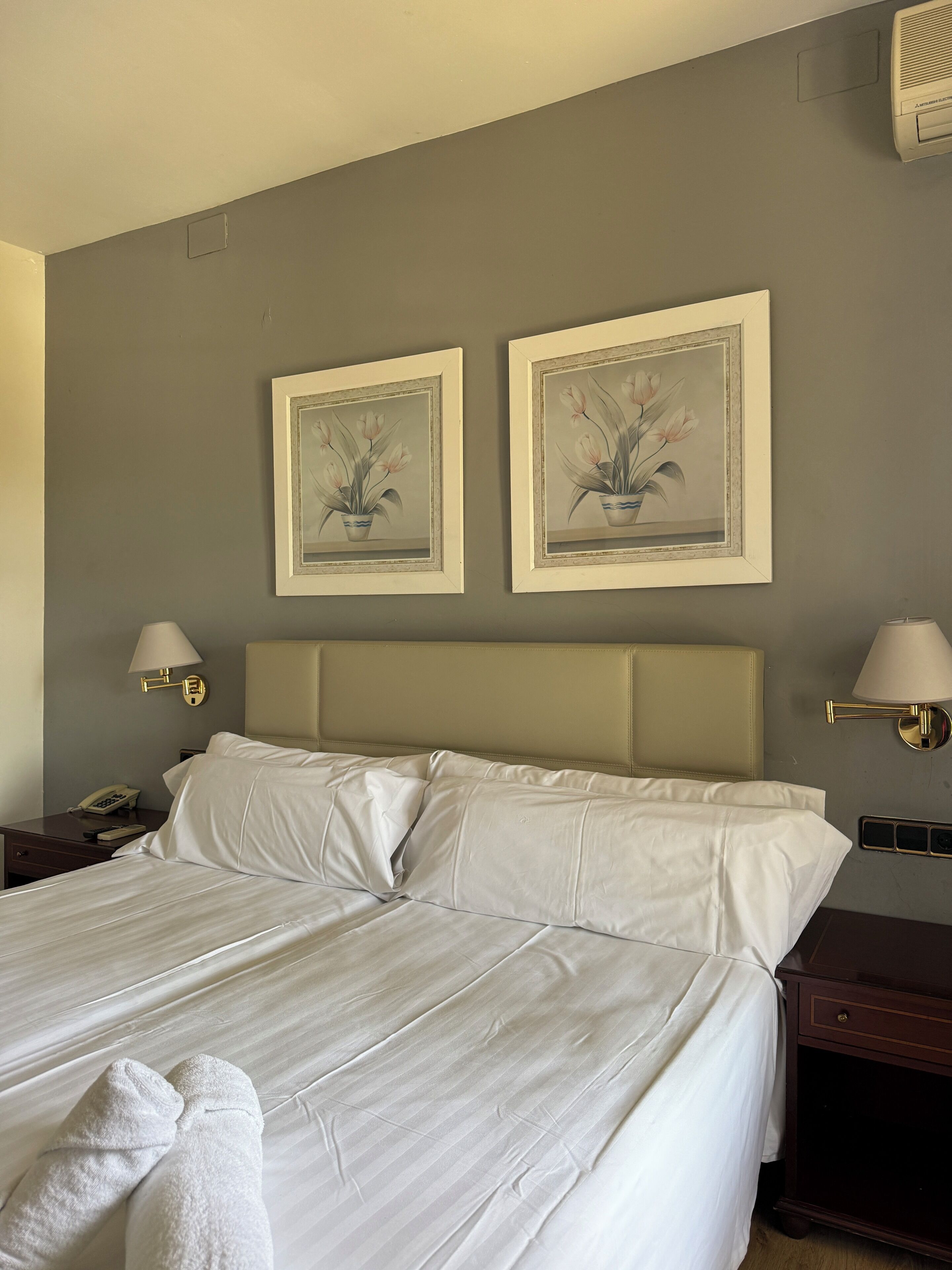 Una habitación de hotel con una cama, dos lámparas de pared, un teléfono y dos cuadros enmarcados con motivos florales.