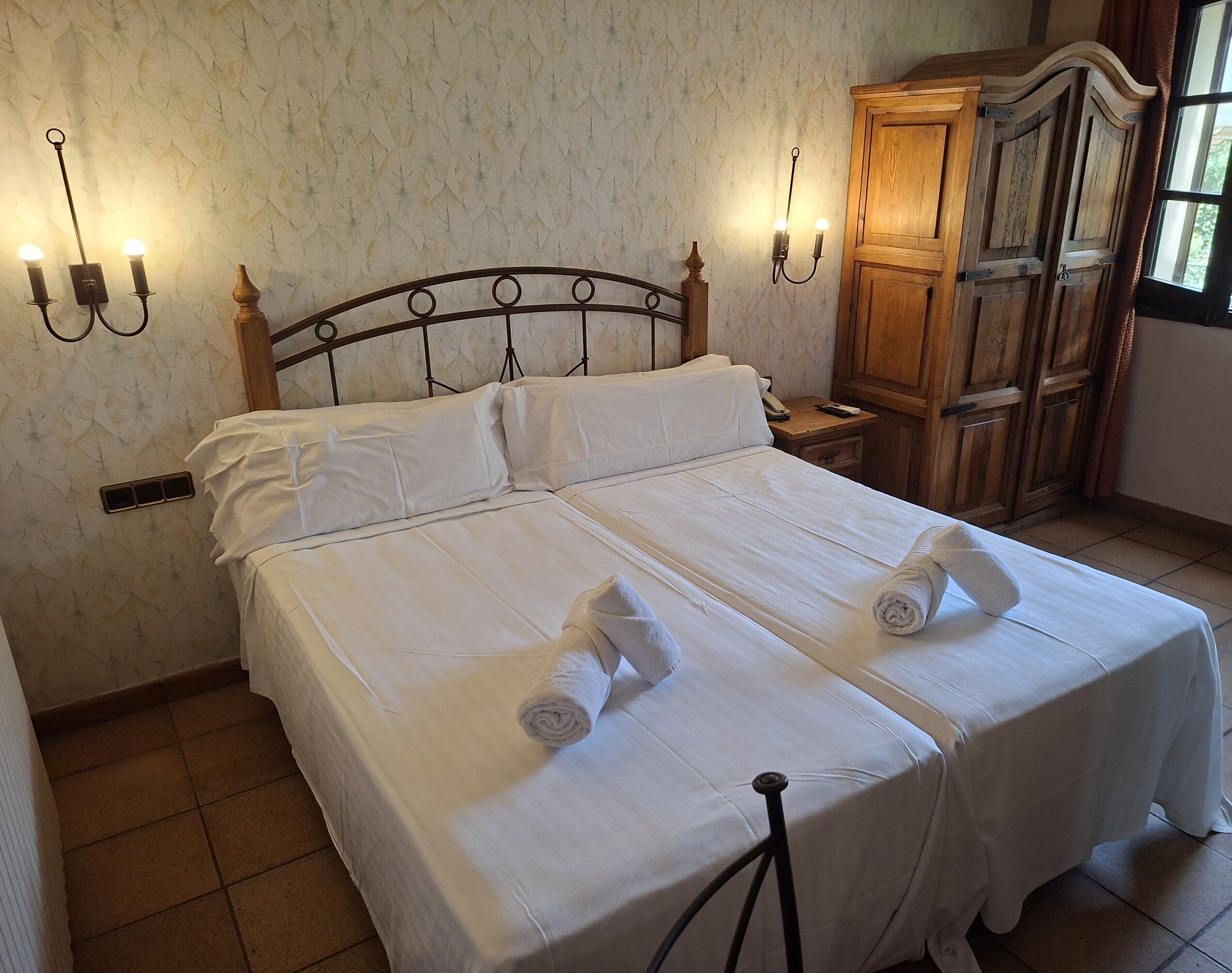 Una cama doble bien tendida con sábanas blancas, dos lámparas de pared, un armario de madera y una ventana con cortinas.