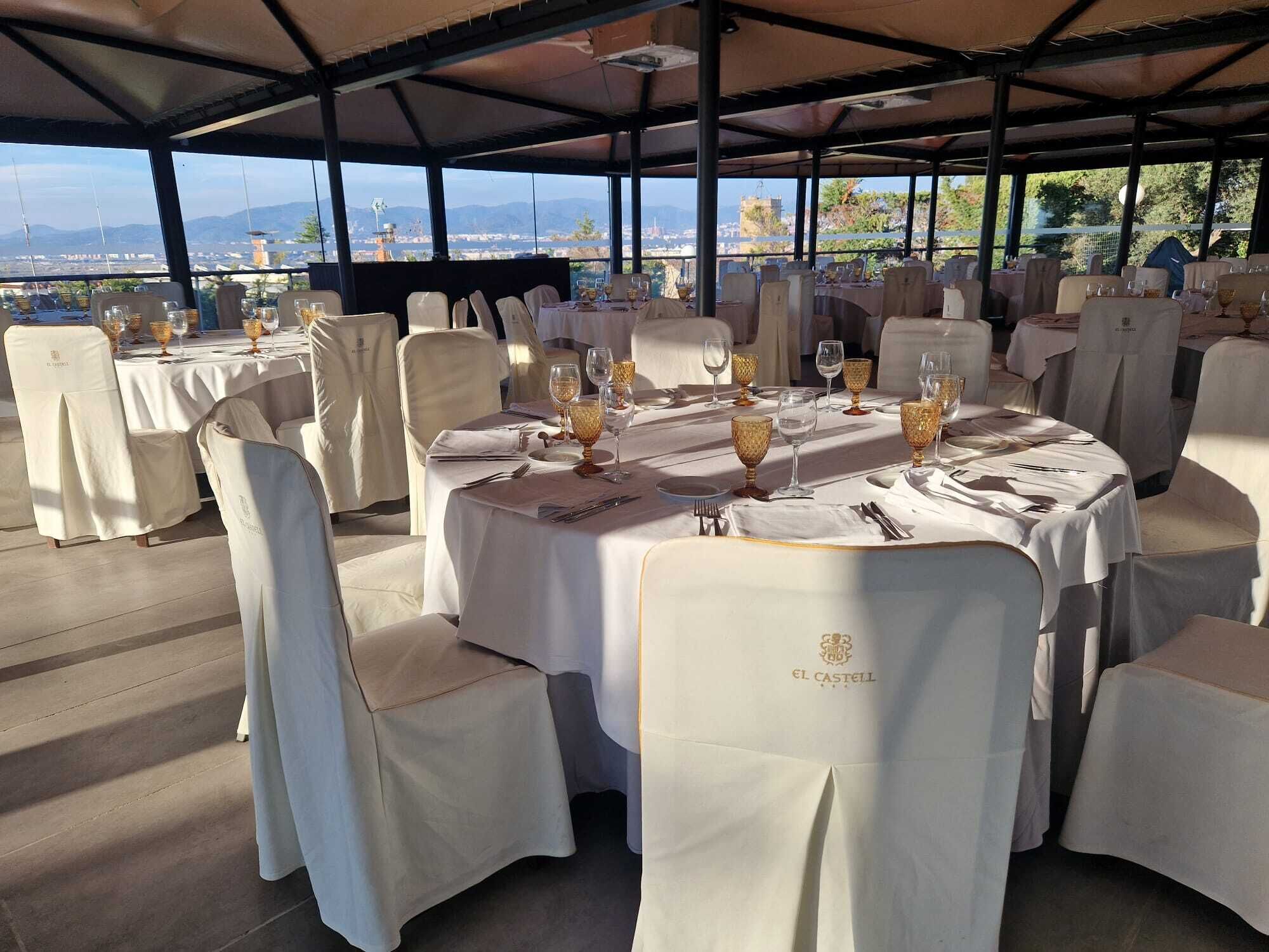 A restaurant with round tables, white tablecloths, and gold-rimmed wine glasses, set against a backdrop of a scenic view.