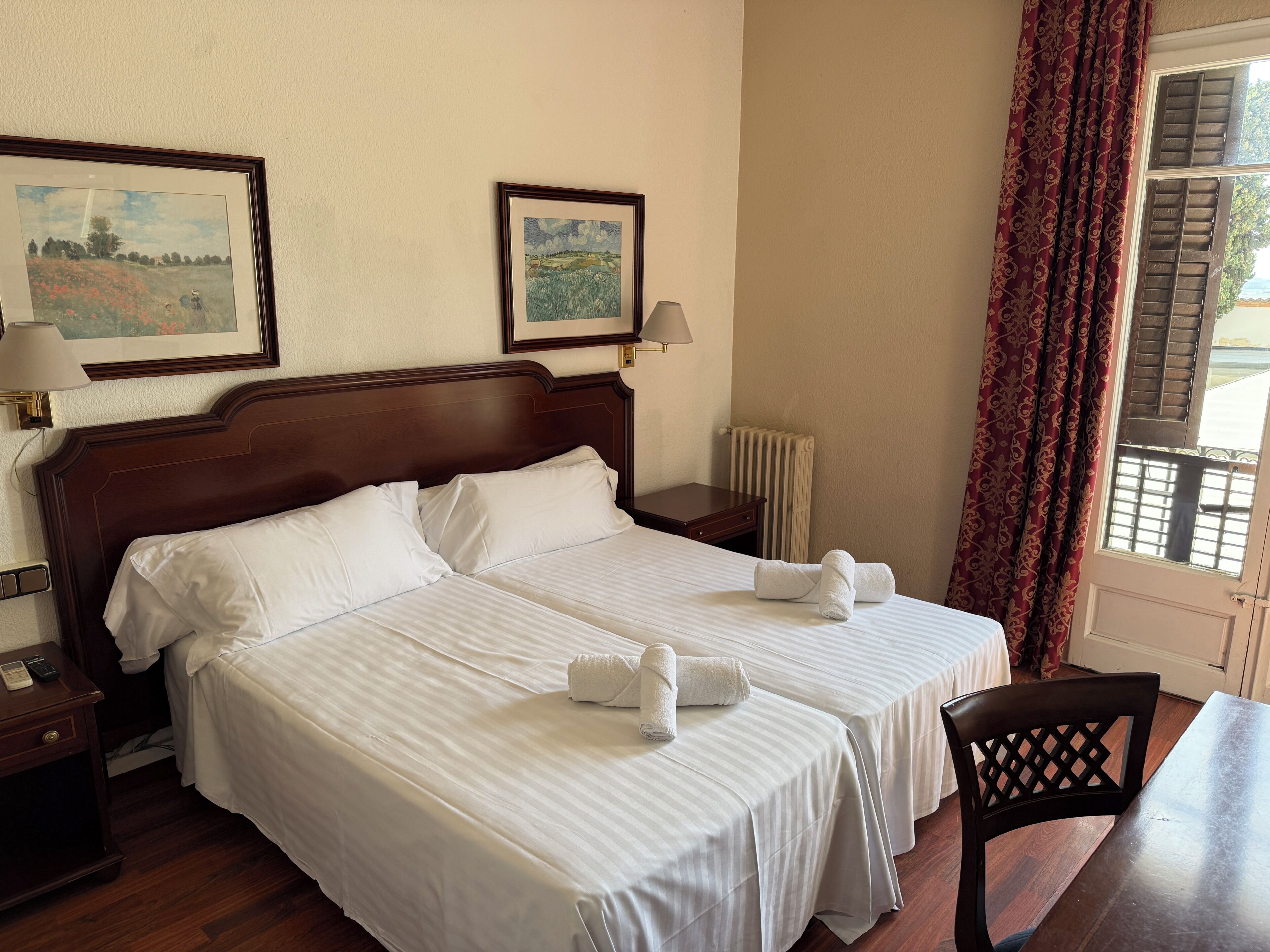 A hotel room with two beds, a desk, a chair, a lamp, and two framed pictures on the wall.