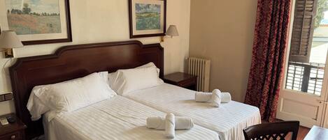 A hotel room with two beds, a desk, a chair, a lamp, and two framed pictures on the wall.