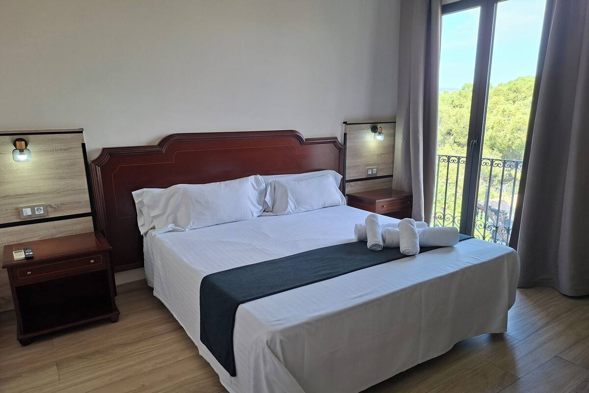 A hotel room with a large bed, wooden headboard, two bedside tables, a nightstand, a window with a view, and a balcony.