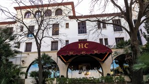 A building with a red awning and the letters 'HC' on it, surrounded by trees and a well-maintained lawn. - Hotel El Castell (Sant Boi de Llobregat)