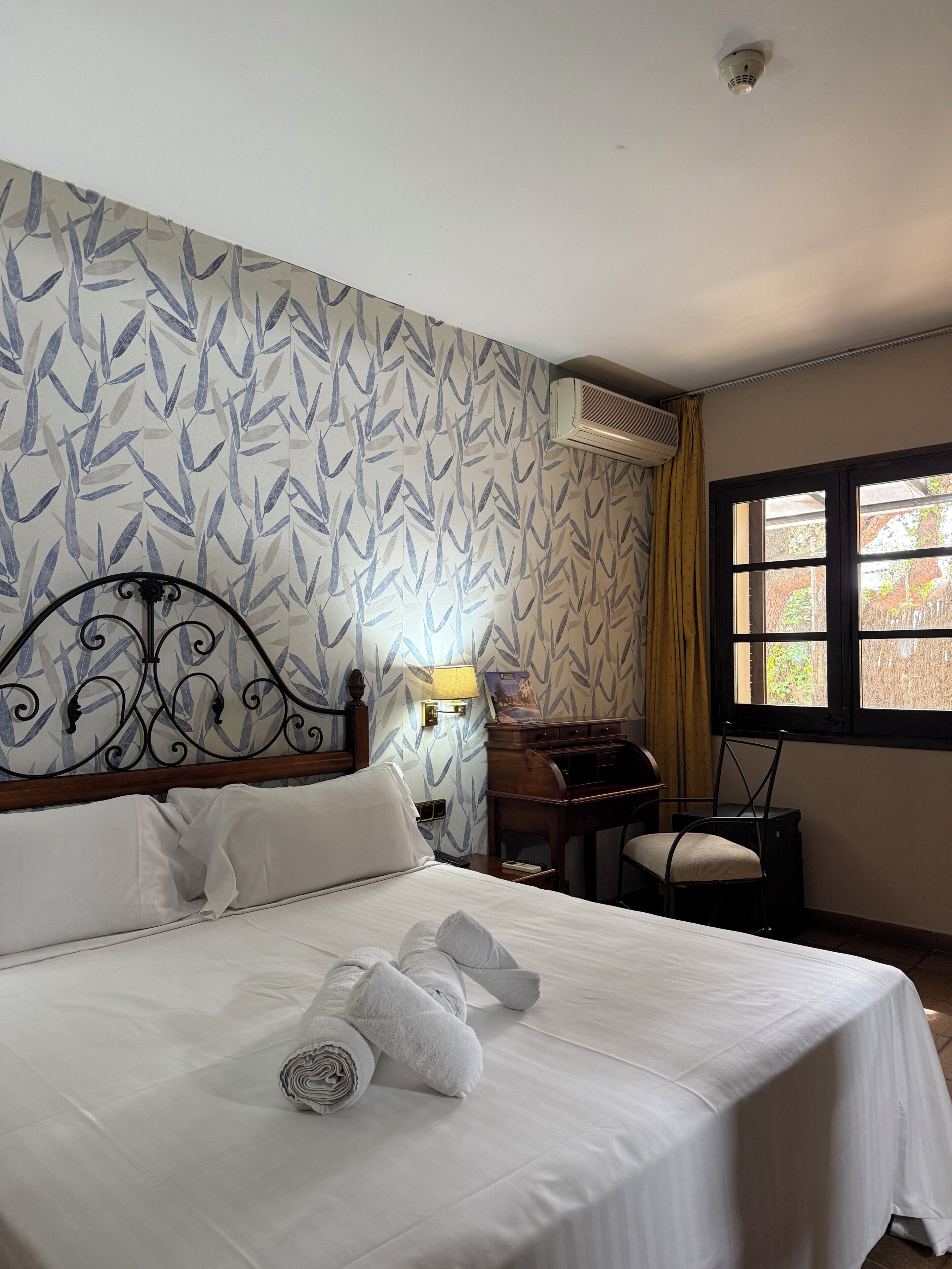 A hotel room with a bed, a desk, a chair, a lamp, and a window with a view of trees.
