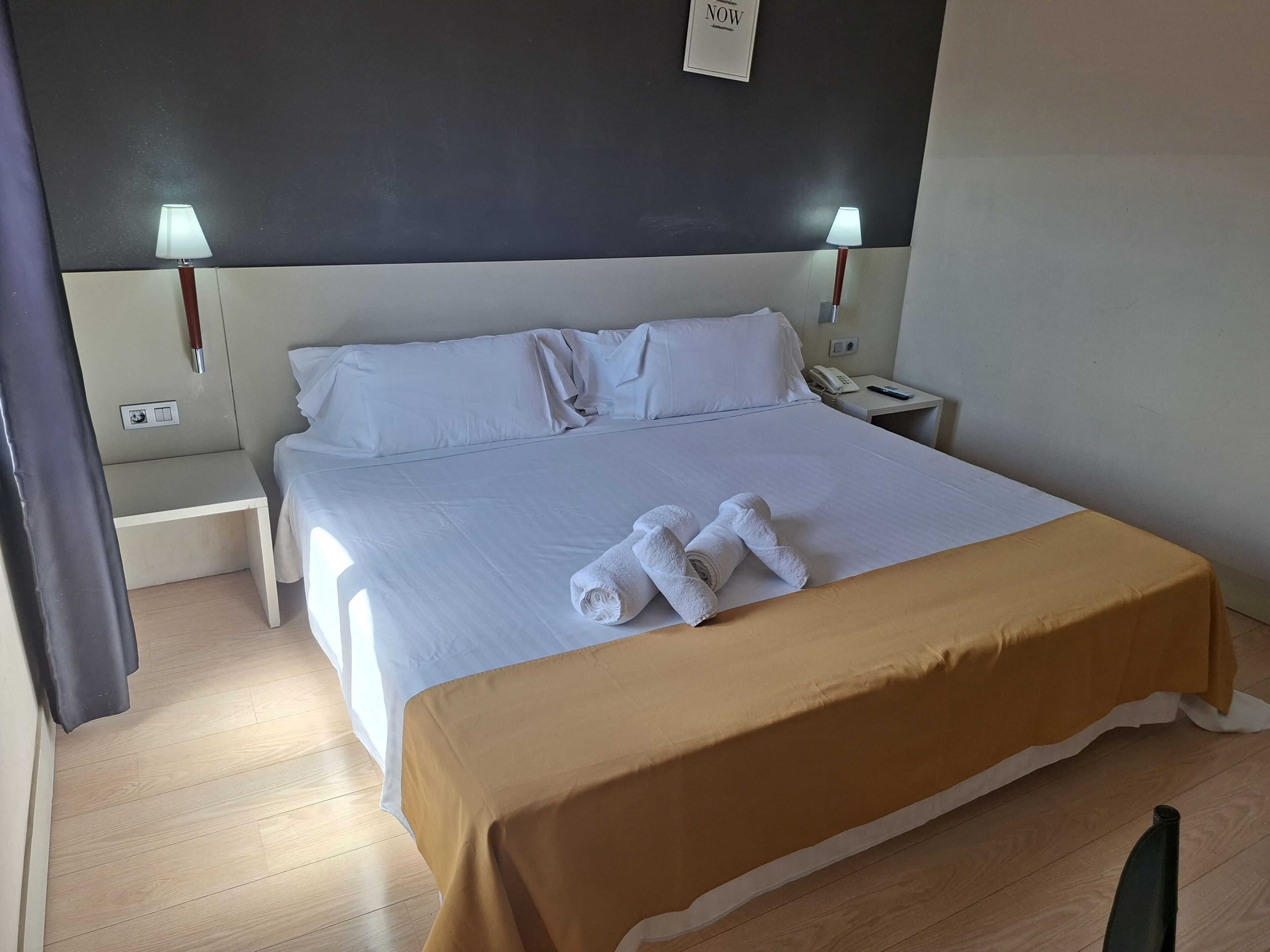 A hotel room with a bed, two bedside lamps, a nightstand with a phone, and a wall-mounted light switch.