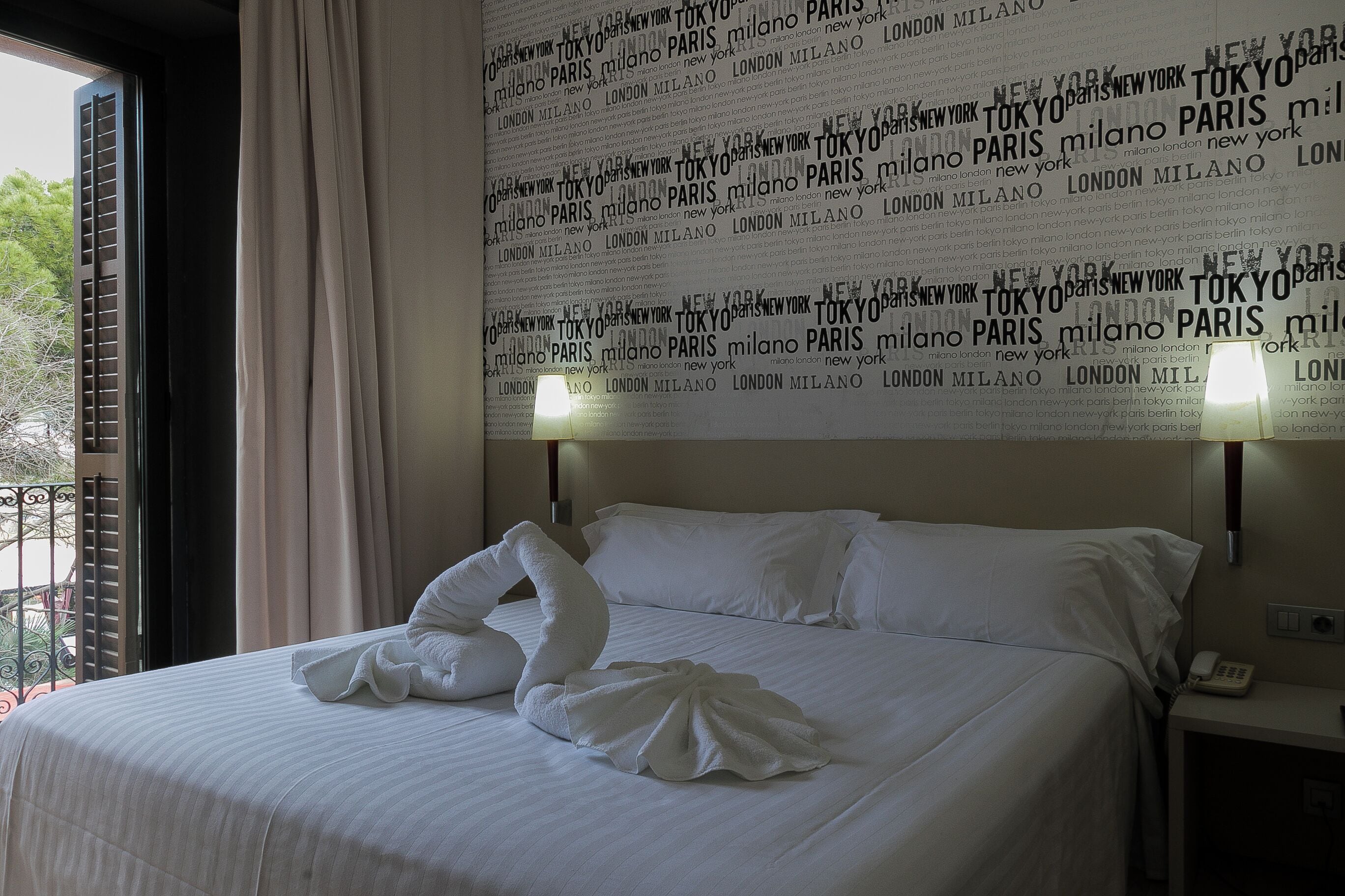 Habitación de hotel con cama, pared con nombres de ciudades, ventana y teléfono en la mesita de noche.