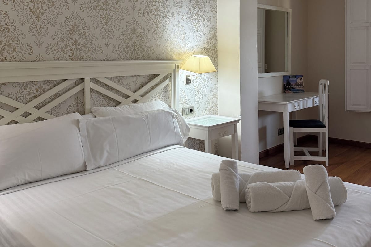A hotel room with a bed, a desk, a chair, a lamp, and a mirror.