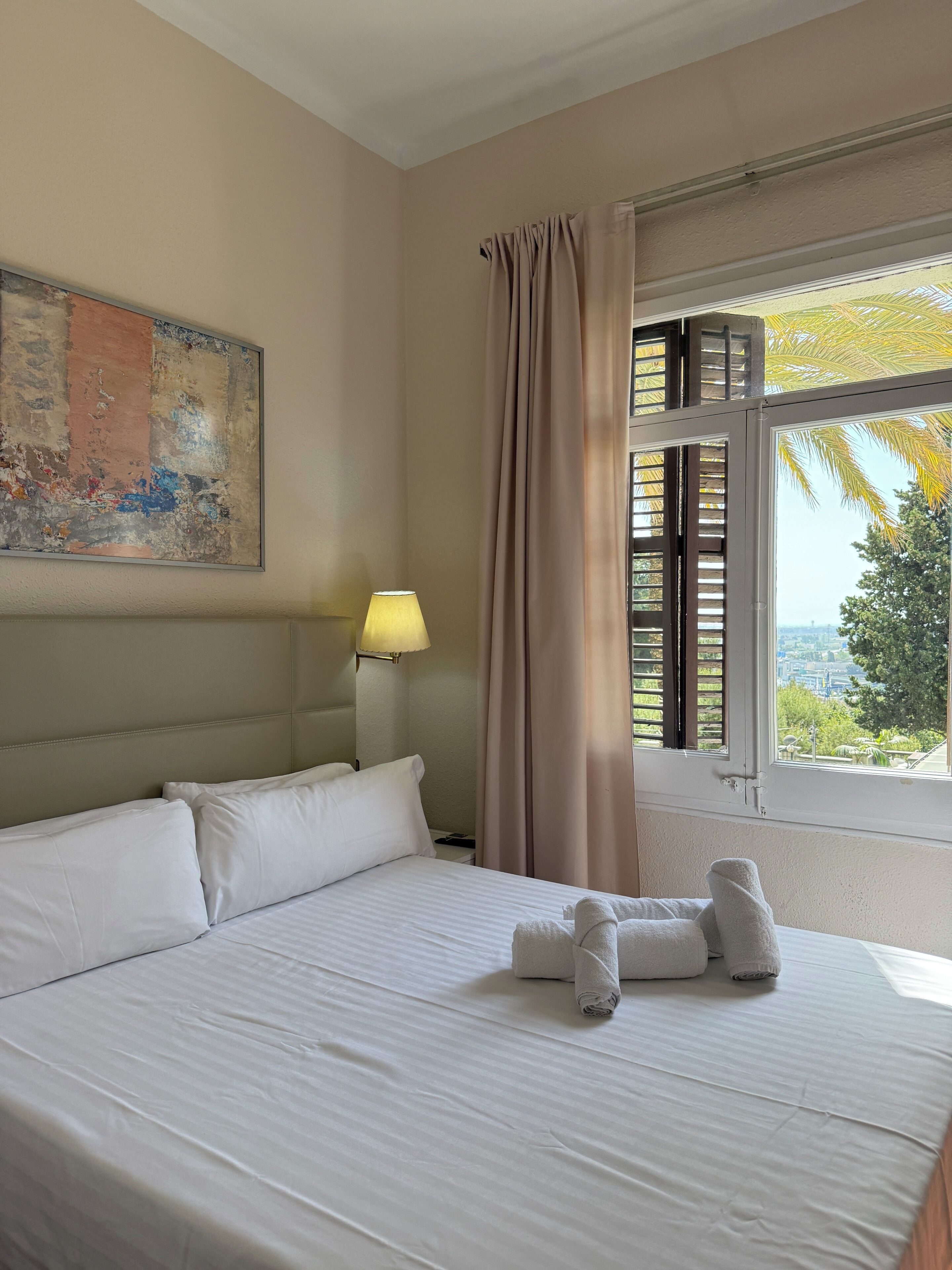A hotel room with a bed, a lamp, a window with curtains, and a painting on the wall.