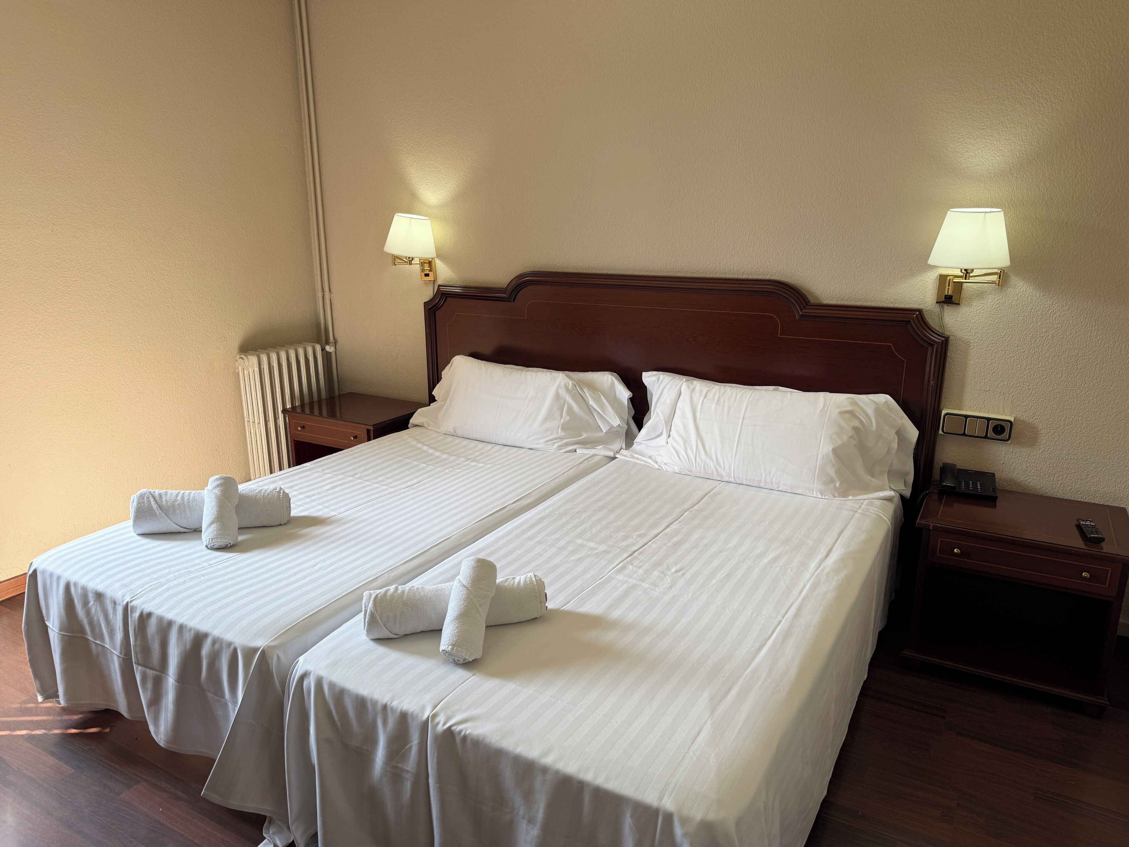 Une chambre d’hôtel avec deux lits, chacun équipé de draps et de serviettes blancs, de têtes de lit en bois, de tables de chevet avec des lampes et d’un radiateur.
