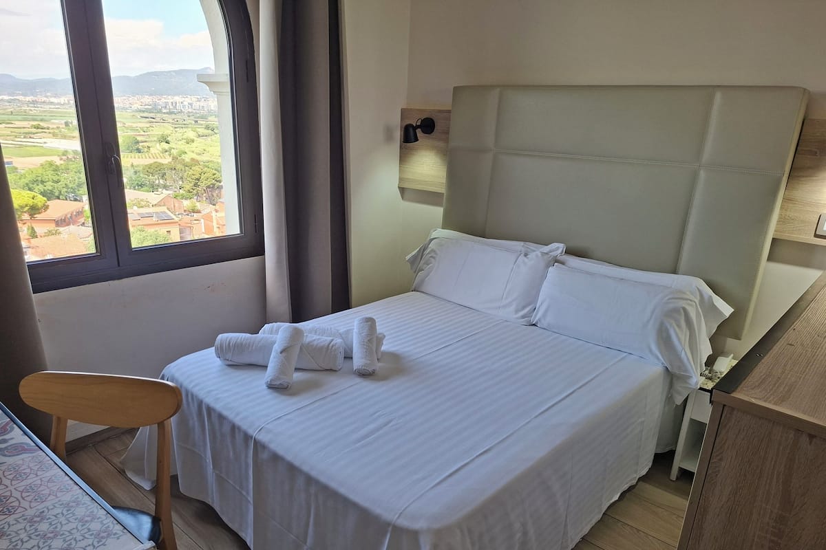 A hotel room with a bed, a chair, a desk, and a view of the countryside.