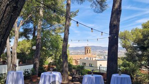 An outdoor dining area with tables set for a meal, surrounded by trees and overlooking a cityscape. - Hotel El Castell (Sant Boi de Llobregat)