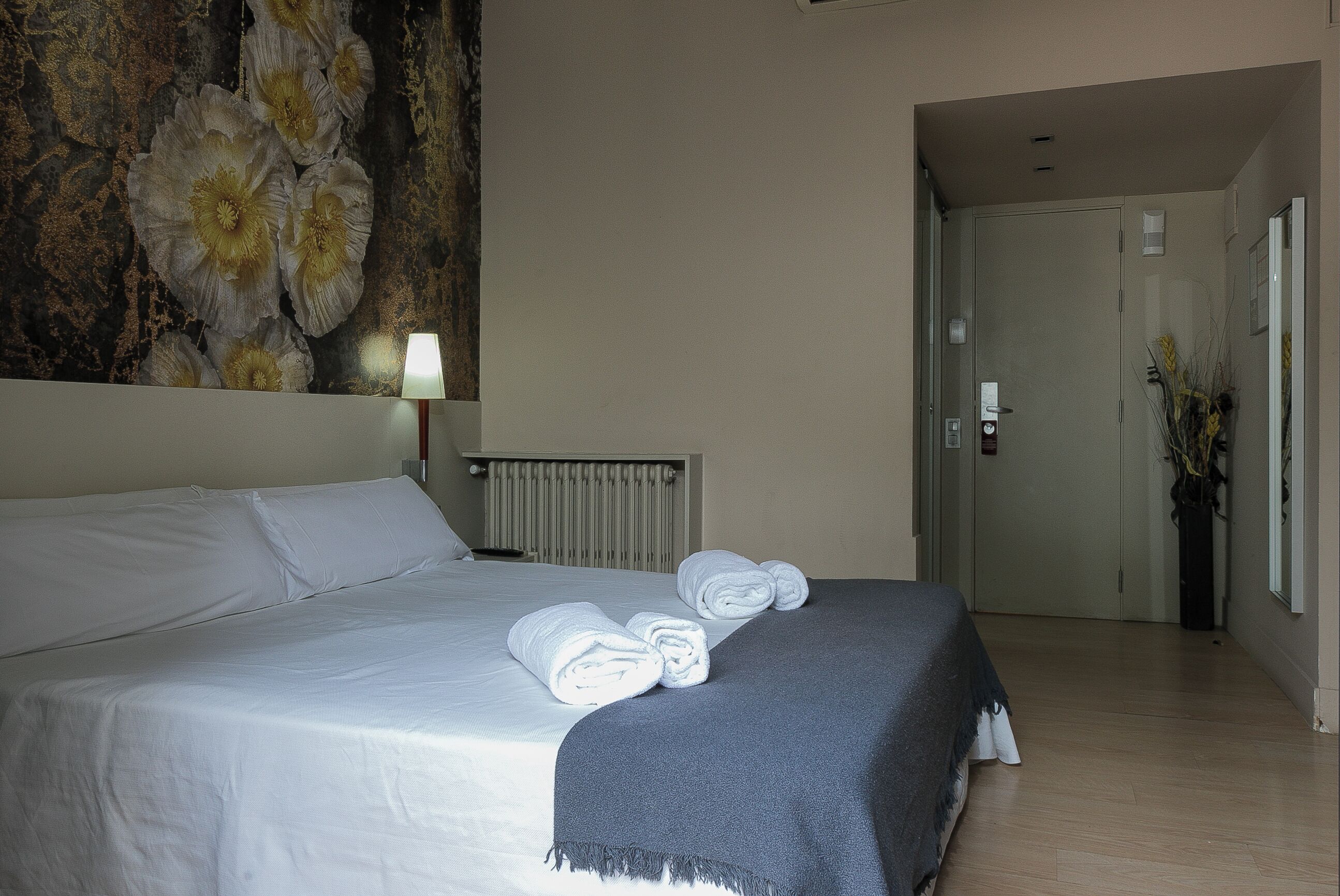 A hotel room with a bed, a wall with a floral mural, a radiator, a door, and a vase with dried plants.