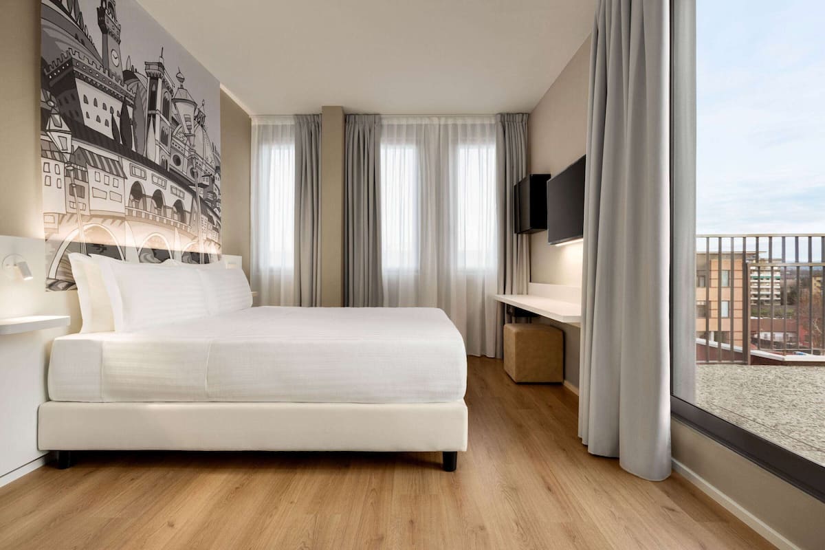 A modern hotel room with a large bed, a desk, a flat-screen TV, and a balcony with a city view.