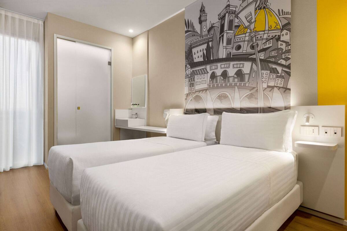 A hotel room with two beds, a bathroom with a mirror, and a wall with a cityscape mural.