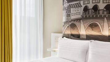 Habitación de hotel con cama, cabecera blanca, lámpara de pared y ventana con cortinas de gasa.