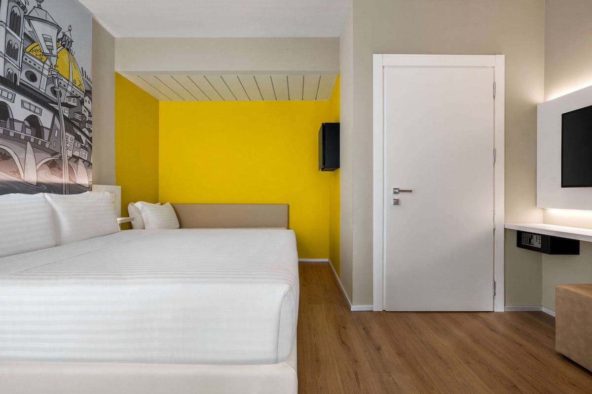 A hotel room with a large bed, a yellow accent wall, a white door, and a mounted TV.