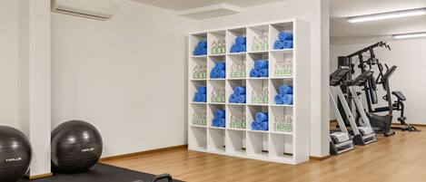 A gym with exercise equipment, a black mat, and shelves with rolled-up towels and water bottles.