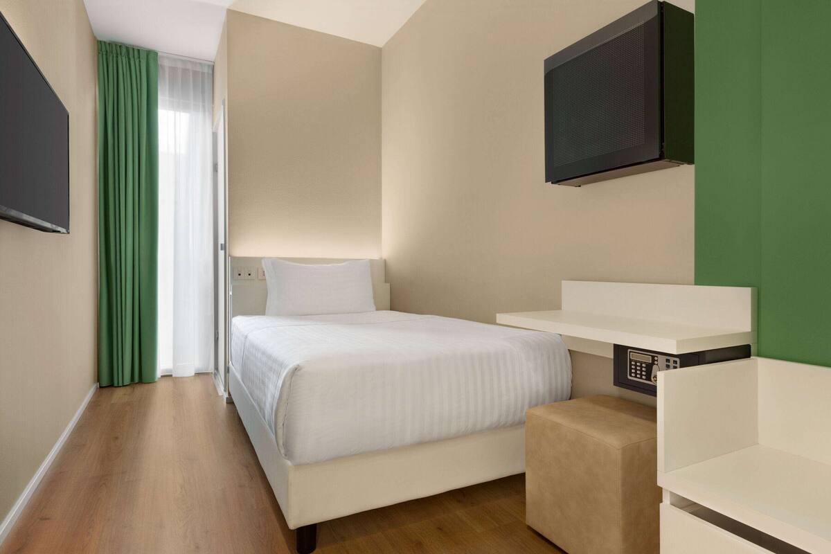 A hotel room with a bed, a TV mounted on the wall, a small bedside table, and a green curtain.