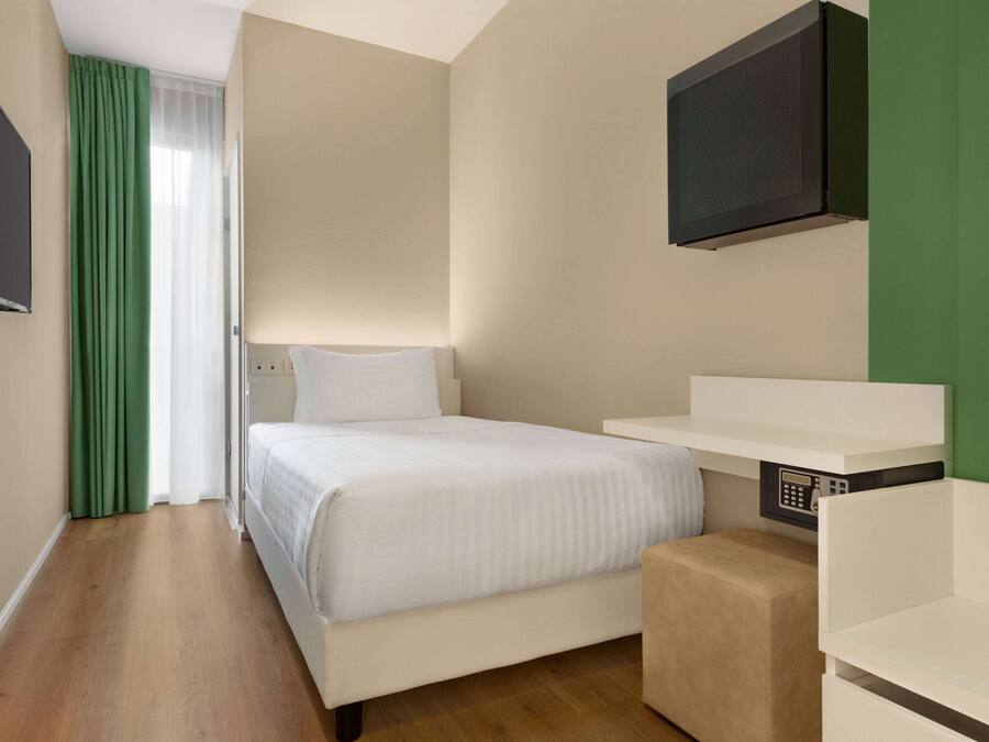 Ein Hotelzimmer mit einem Bett, einem an der Wand montierten Fernseher, einem kleinen Nachttisch und einem grünen Vorhang.
