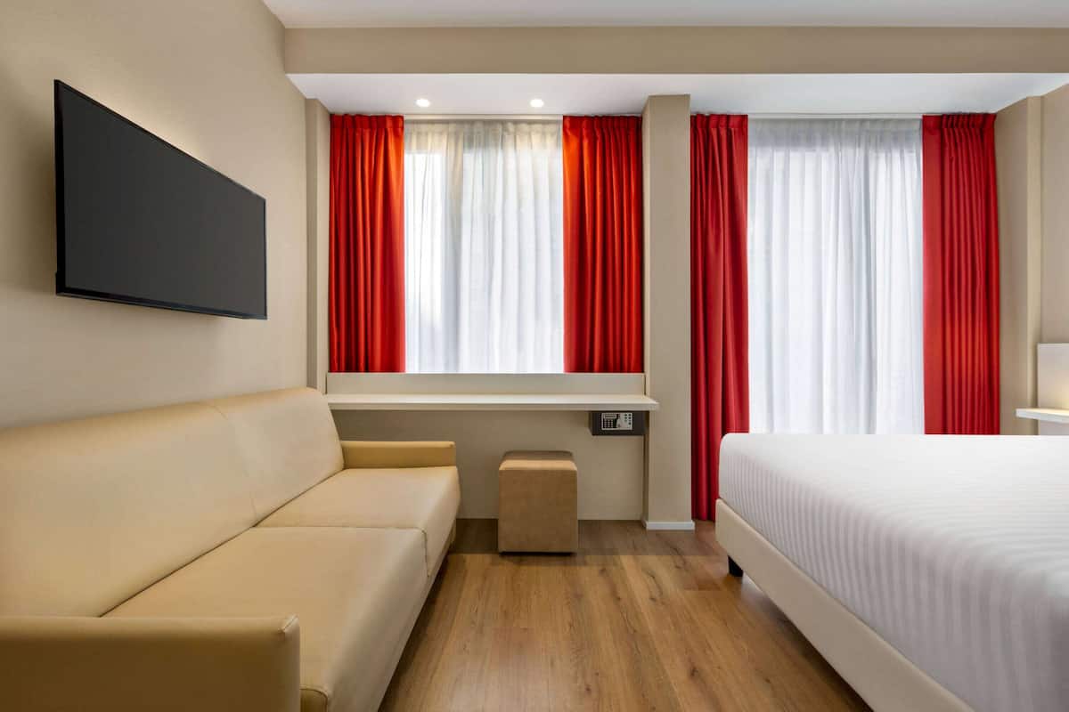A hotel room with a sofa, a bed, a TV mounted on the wall, and a window with red curtains.