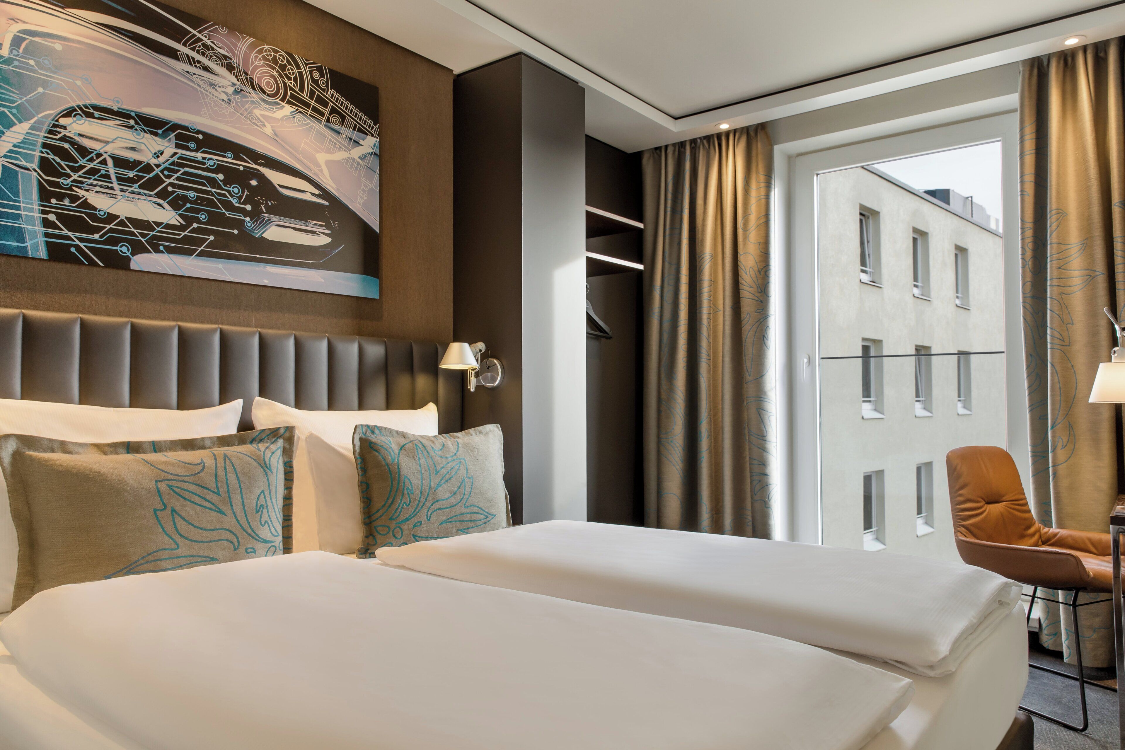 A hotel room with a large bed, a headboard, a seating area with a chair, and a view of buildings through the window.