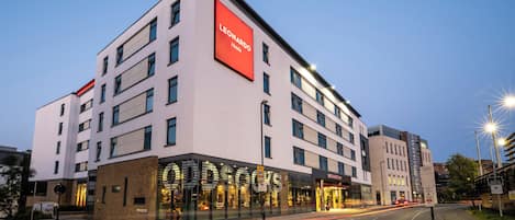A modern hotel building with a prominent 'Leonardo Hotels' sign, large windows, and a well-lit entrance.