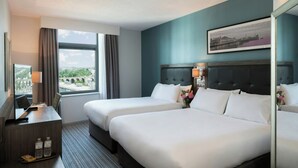 A hotel room with two beds, a desk with a lamp, a window with curtains, and a framed picture on the wall.