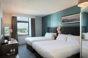 A hotel room with two beds, a desk with a lamp, a window with curtains, and a framed picture on the wall.