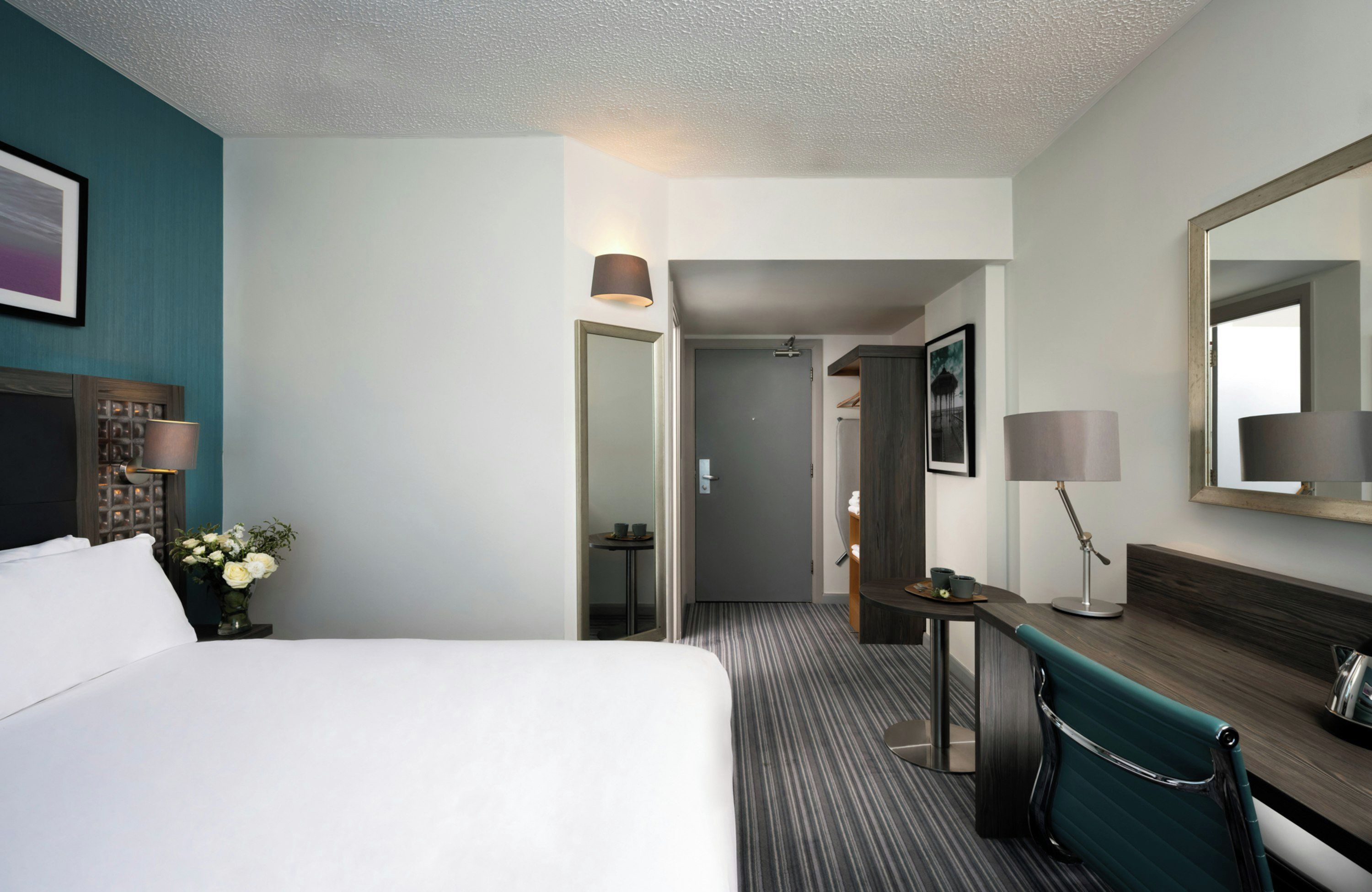 A hotel room with a bed, a desk, a chair, a mirror, and a door leading to another room.