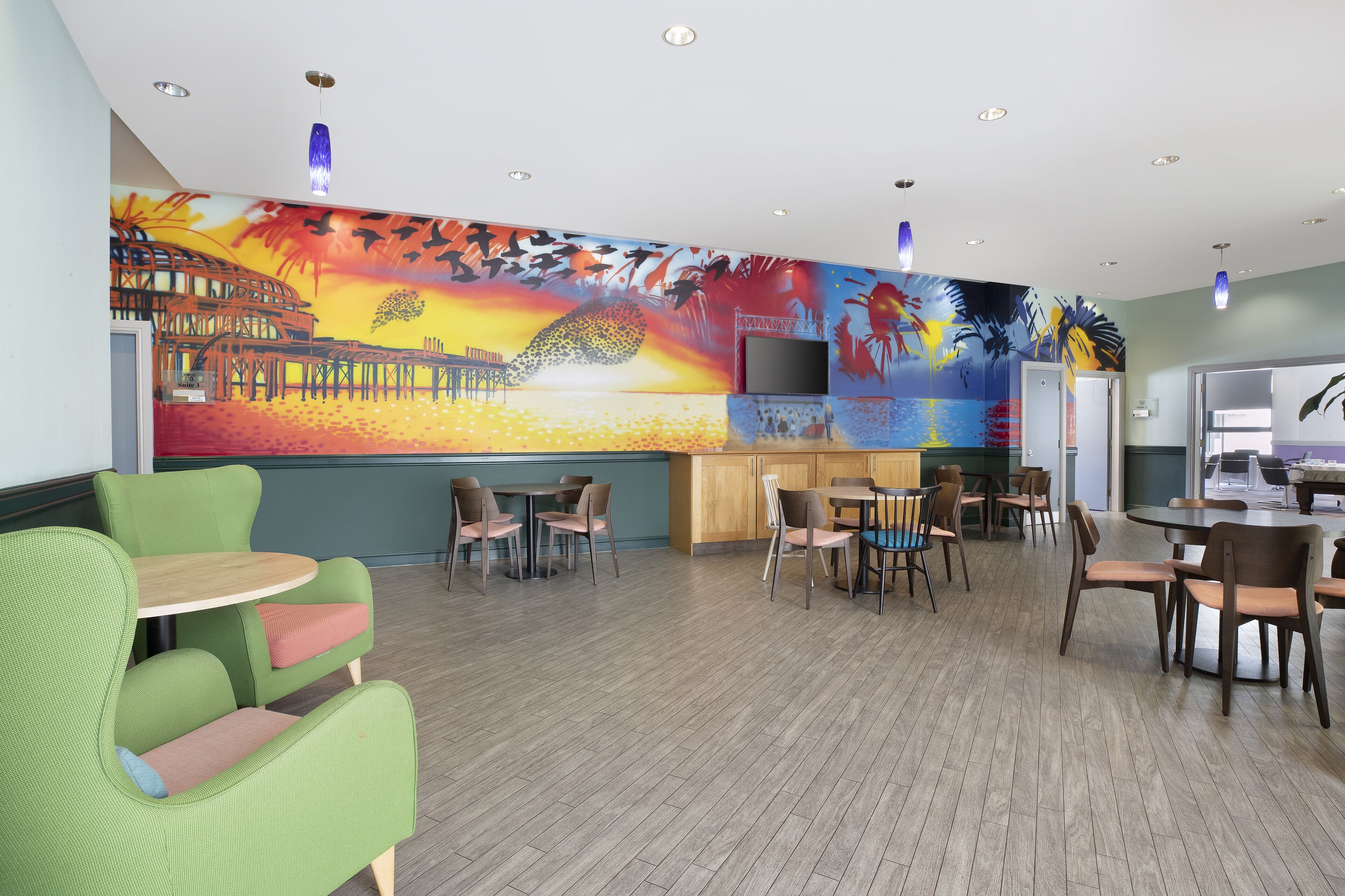 A spacious lounge area with multiple seating options including green chairs and tables, a large mural on the wall, and a flat-screen TV mounted on the wall.