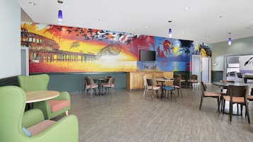A spacious lounge area with multiple seating options including green chairs and tables, a large mural on the wall, and a flat-screen TV mounted on the wall.