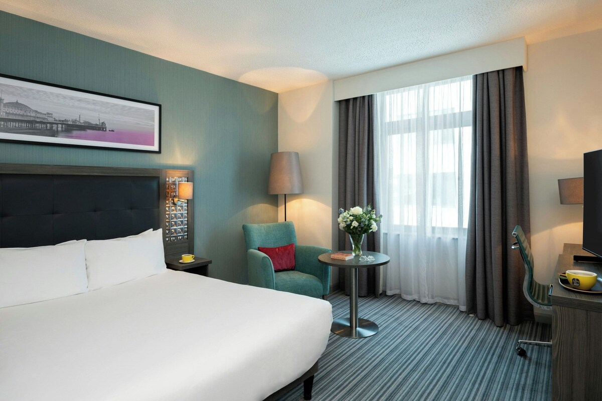 A hotel room with a bed, a chair, a table with a vase of flowers, a lamp, and a television.