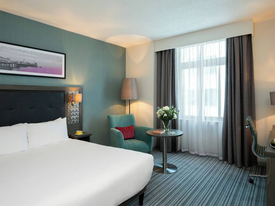 A hotel room with a bed, a chair, a table with a vase of flowers, a lamp, and a television.