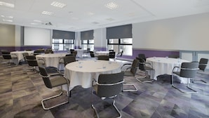 A conference room with multiple round tables, each surrounded by chairs. The room has a carpeted floor and large windows allowing natural light.