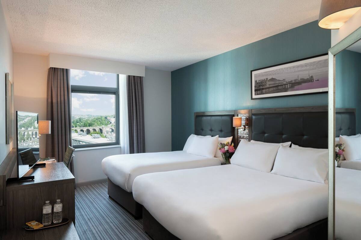 A hotel room with two beds, a desk with a lamp, a large window with curtains, and a framed picture on the wall.