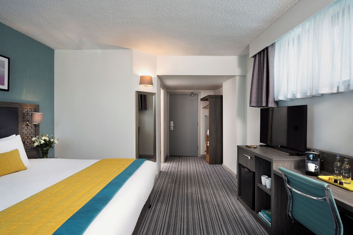 A hotel room with a bed, a desk, a TV, and a bathroom visible through an open door.