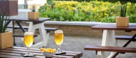 Two tables with beer glasses and a bowl of fries, set outdoors with a hedge and buildings in the background.